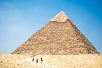 People riding a camel near a pyramid under a blue sky