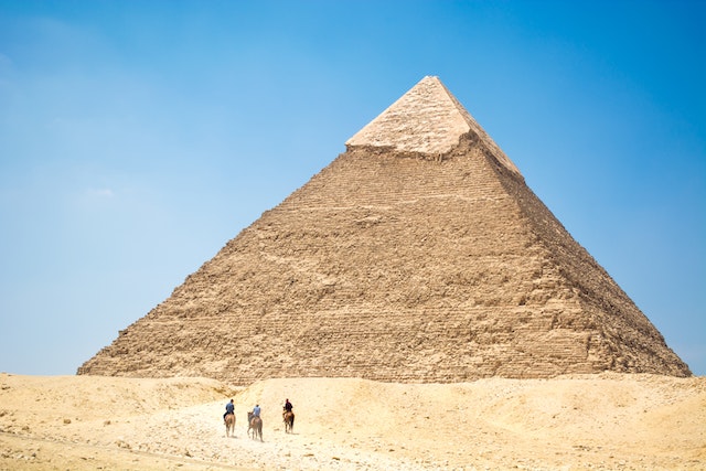 People riding a camel near a pyramid under a blue sky