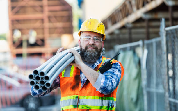 A mature man working at a construction site, carrying pvc pipes on his shoulder. He is wearing a hardhat, safety glasses, and reflective vest. He is working on the renovation of a strip mall.