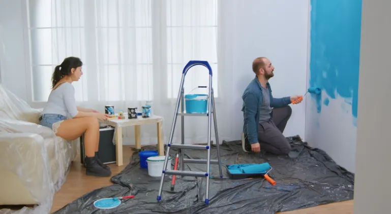 A guy painting a wall of a room while women instructing him